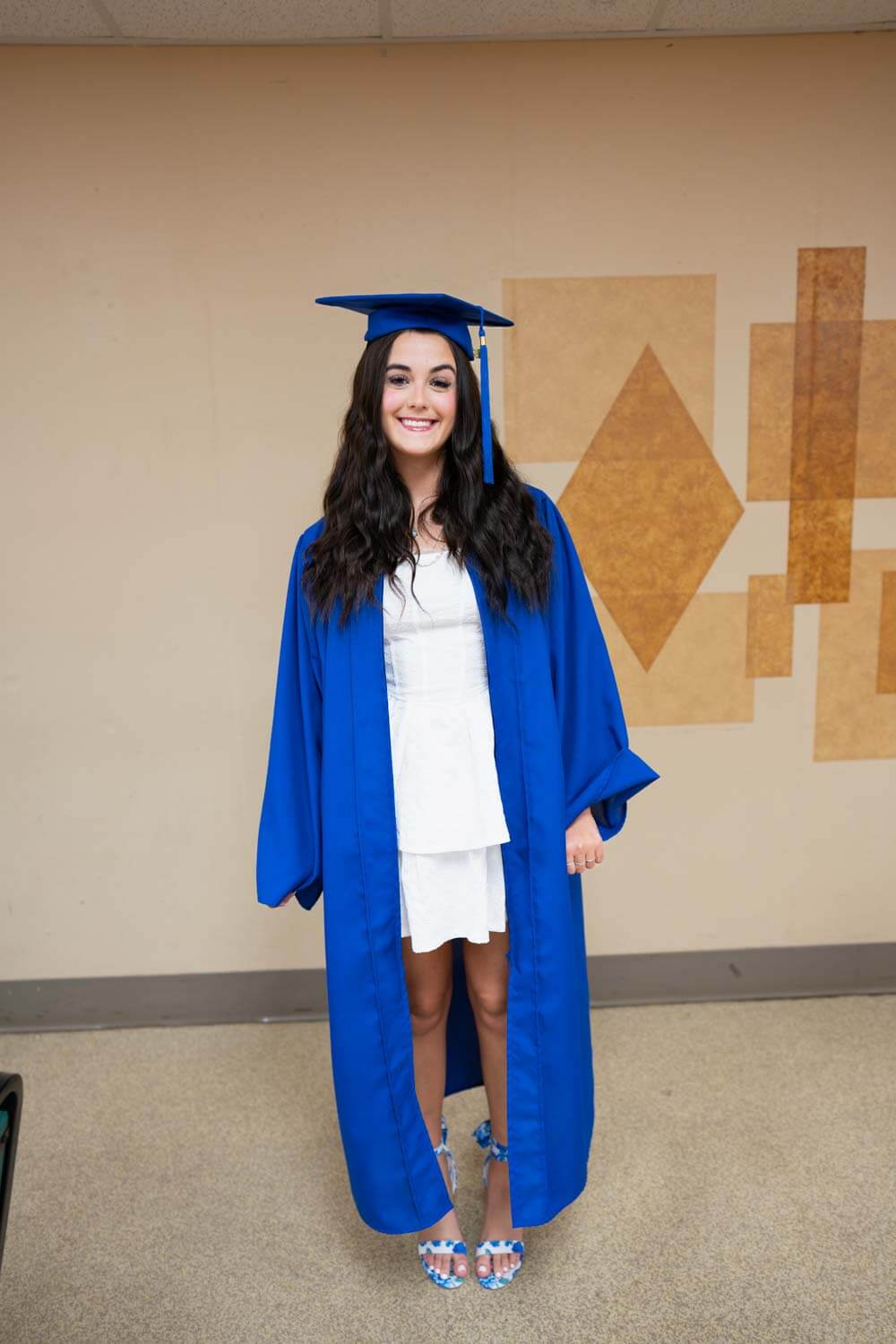 A young woman smiles, wearing a blue graduation cap and gown over a white dress.