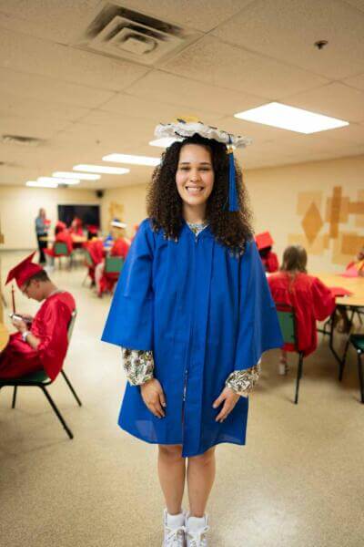 A smiling young woman in a blue graduation gown and decorated cap stands in a classroom.