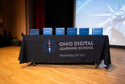 A table with a black cloth bearing the Ohio Digital Learning School logo is topped with neatly arranged blue diplomas