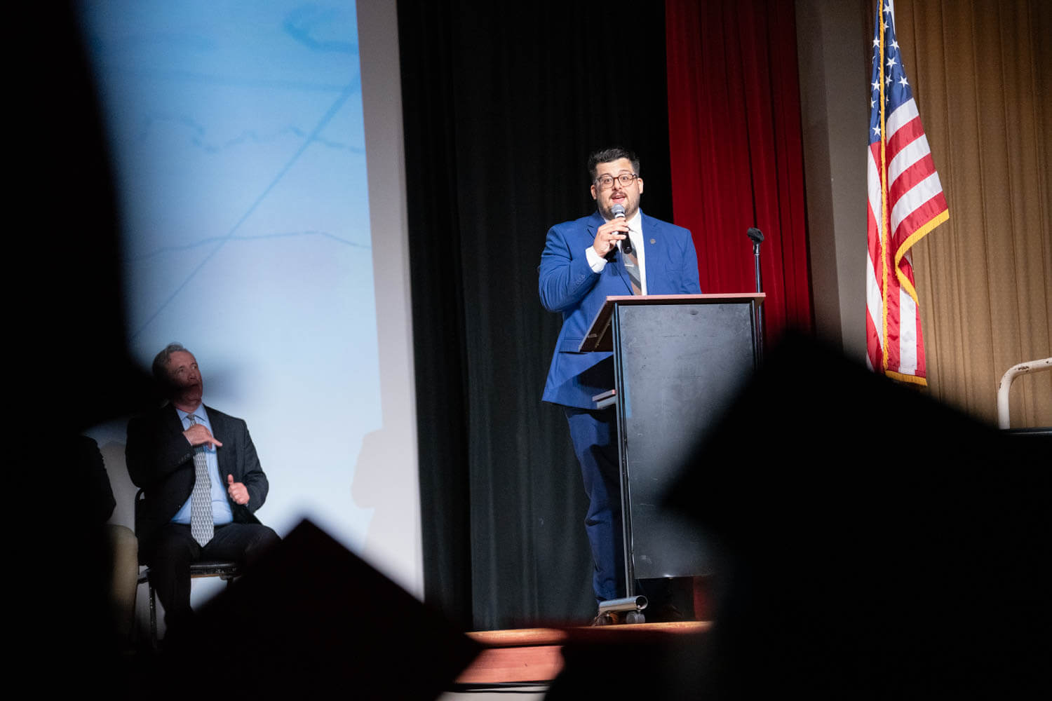 A man in a blue suit speaks at a podium on stage with an American flag beside him.