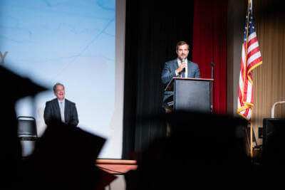 A man speaks at a podium on a stage with an American flag nearby, while another man sits and smiles.