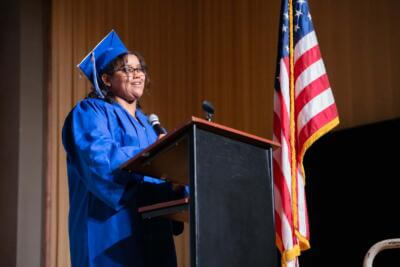 A graduate in a blue cap and gown speaks confidently at a podium with a microphone.