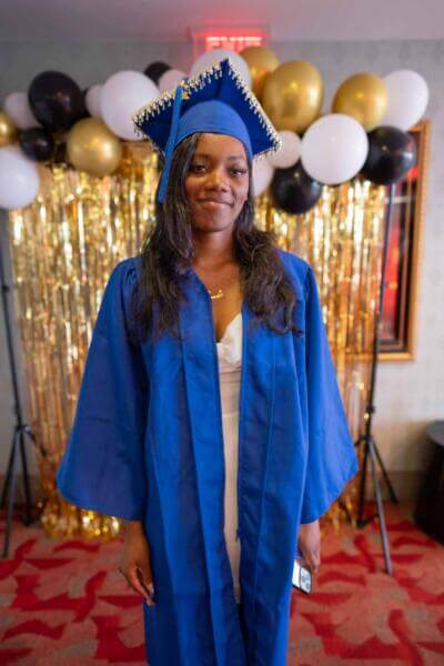 Young woman in blue graduation gown and cap stands smiling in front of festive gold and black balloons and a shimmering backdrop