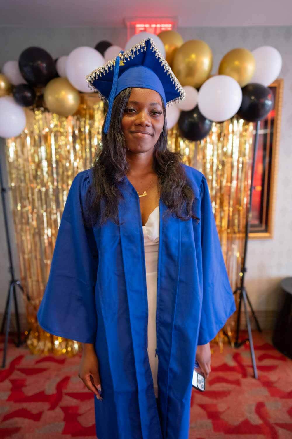 Young woman in blue graduation gown and cap stands smiling in front of festive gold and black balloons and a shimmering backdrop