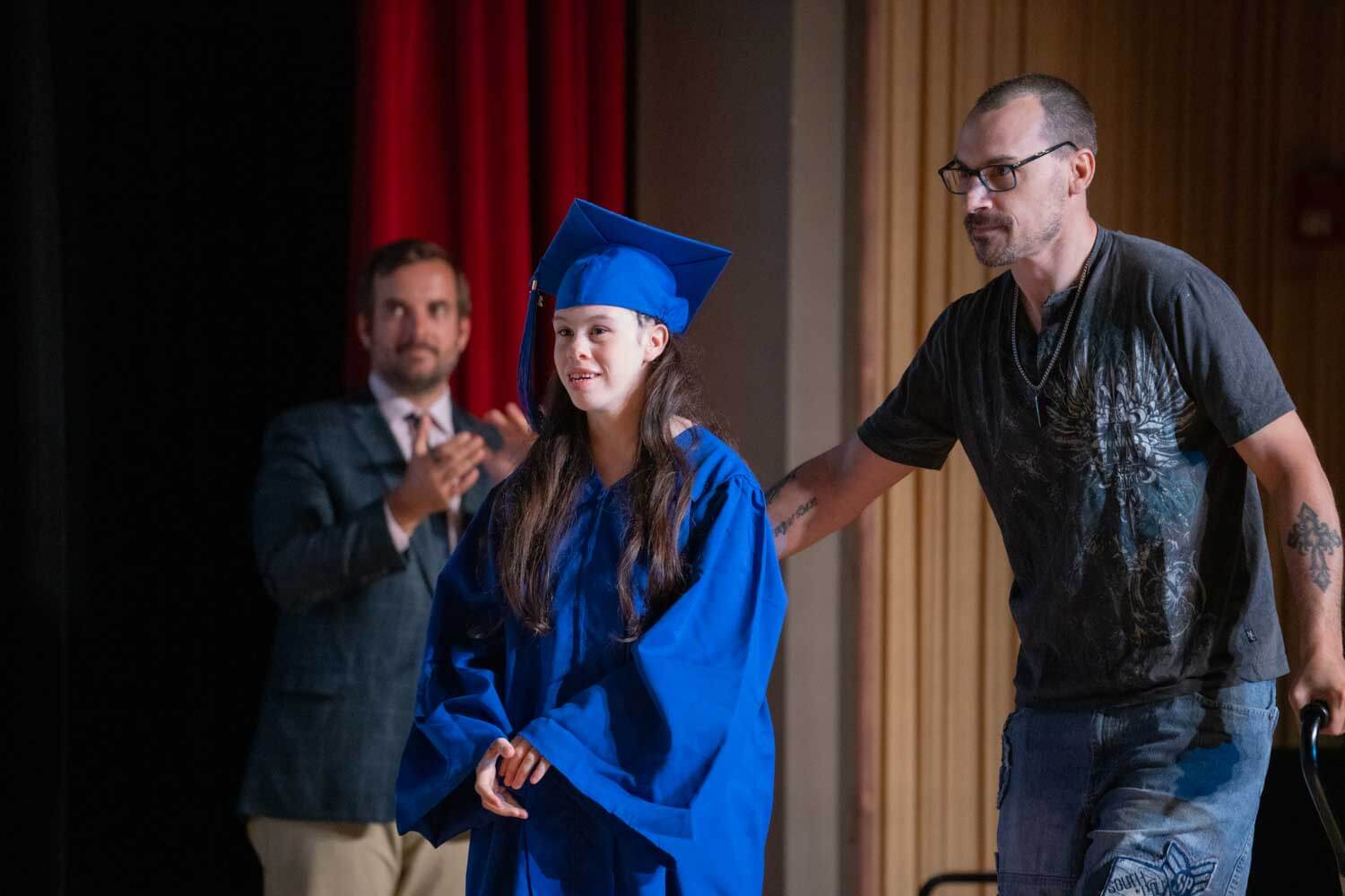 A young woman in a blue graduation gown and cap is guided onstage by a man.