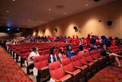 Graduates in red and blue gowns and caps fill auditorium seats, some standing and chatting.