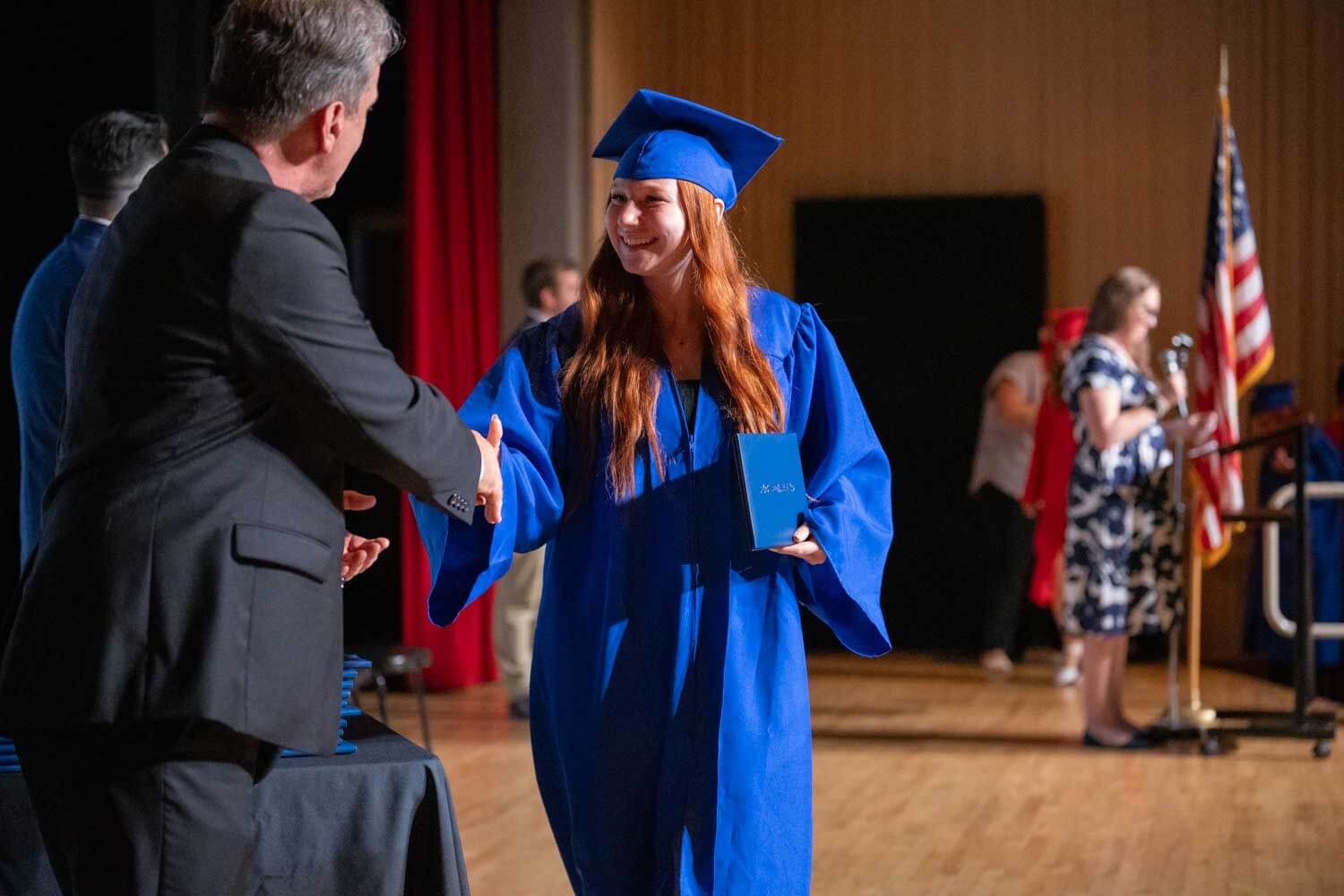 A young woman in a blue cap and gown smiles while shaking hands with a man on stage at a graduation ceremony.