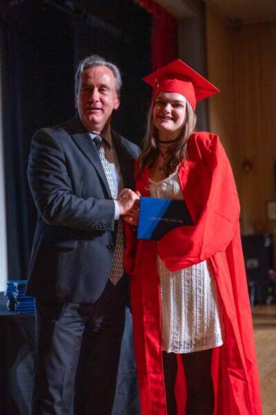 A graduate in a red cap and gown smiles as she shakes hands with a man in a suit on stage, holding a diploma.