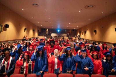 Graduates in red and blue caps and gowns fill an auditorium, adjusting tassels.