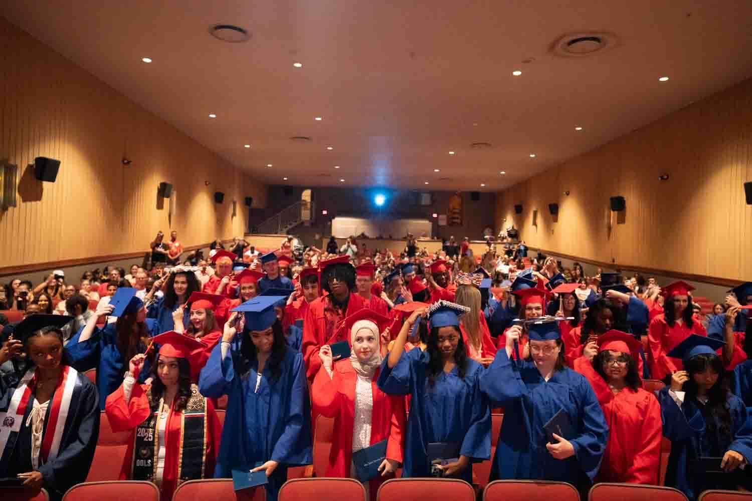 Graduates in red and blue caps and gowns fill an auditorium, adjusting tassels.