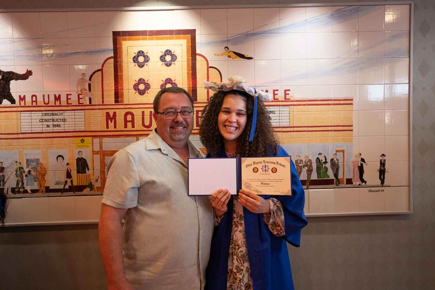 A smiling young woman in a blue graduation gown and cap holds a diploma, standing beside a man.