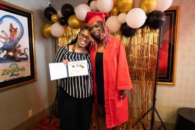 A graduate in a red cap and gown poses with a woman holding a certificate.