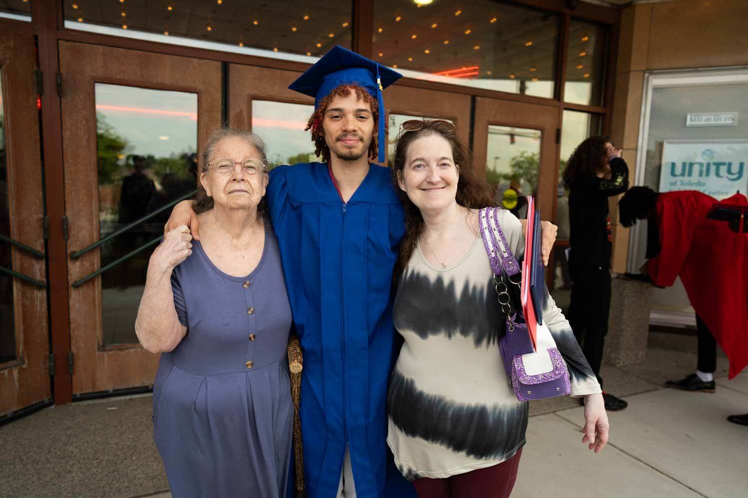 Graduate in a blue cap and gown stands smiling between an elderly woman and a middle-aged woman