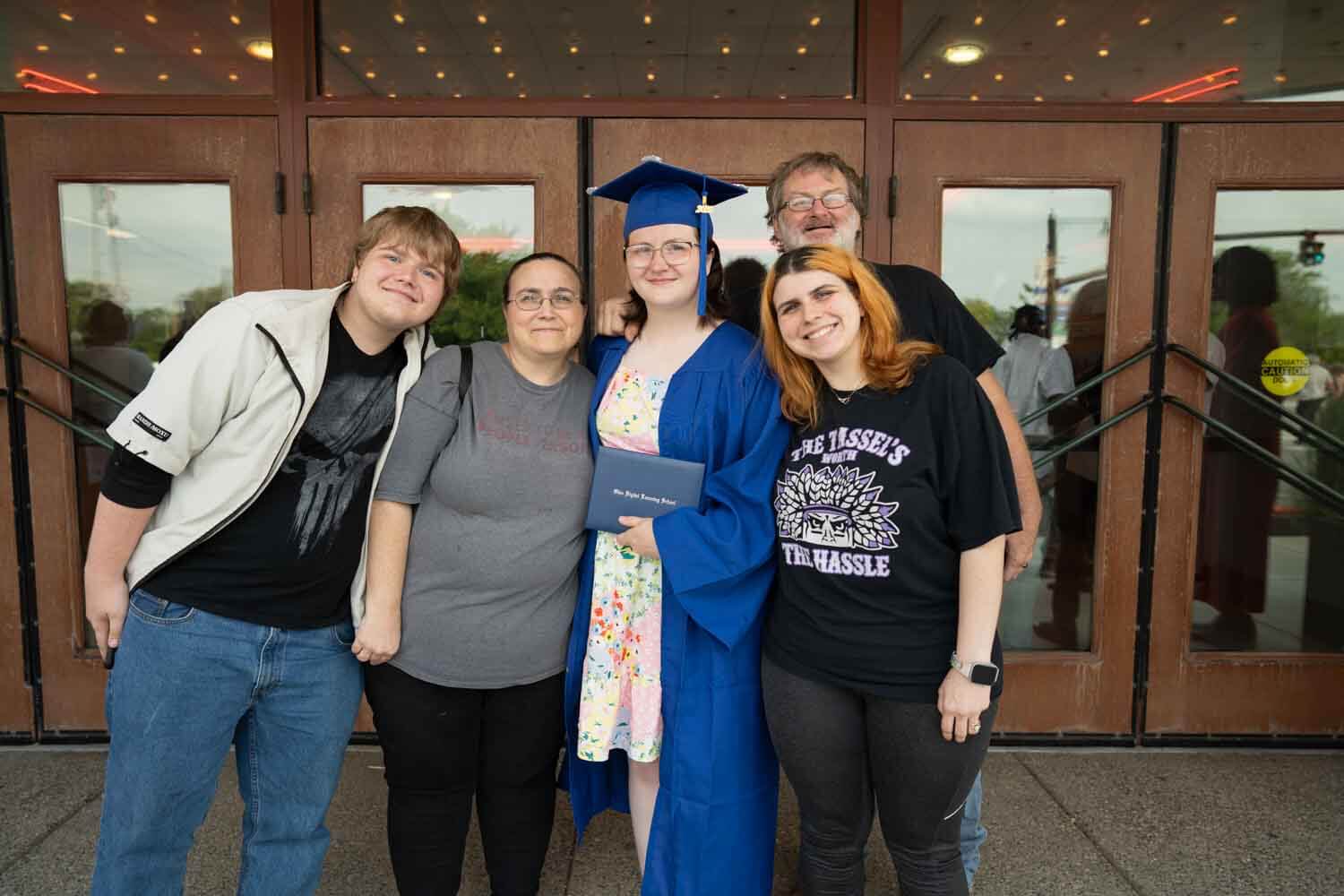 A graduate in a blue cap and gown holds a diploma, surrounded by four smiling people.