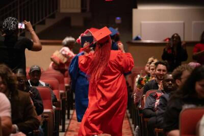 A graduate in a red cap and gown dances joyfully down the aisle of an auditorium