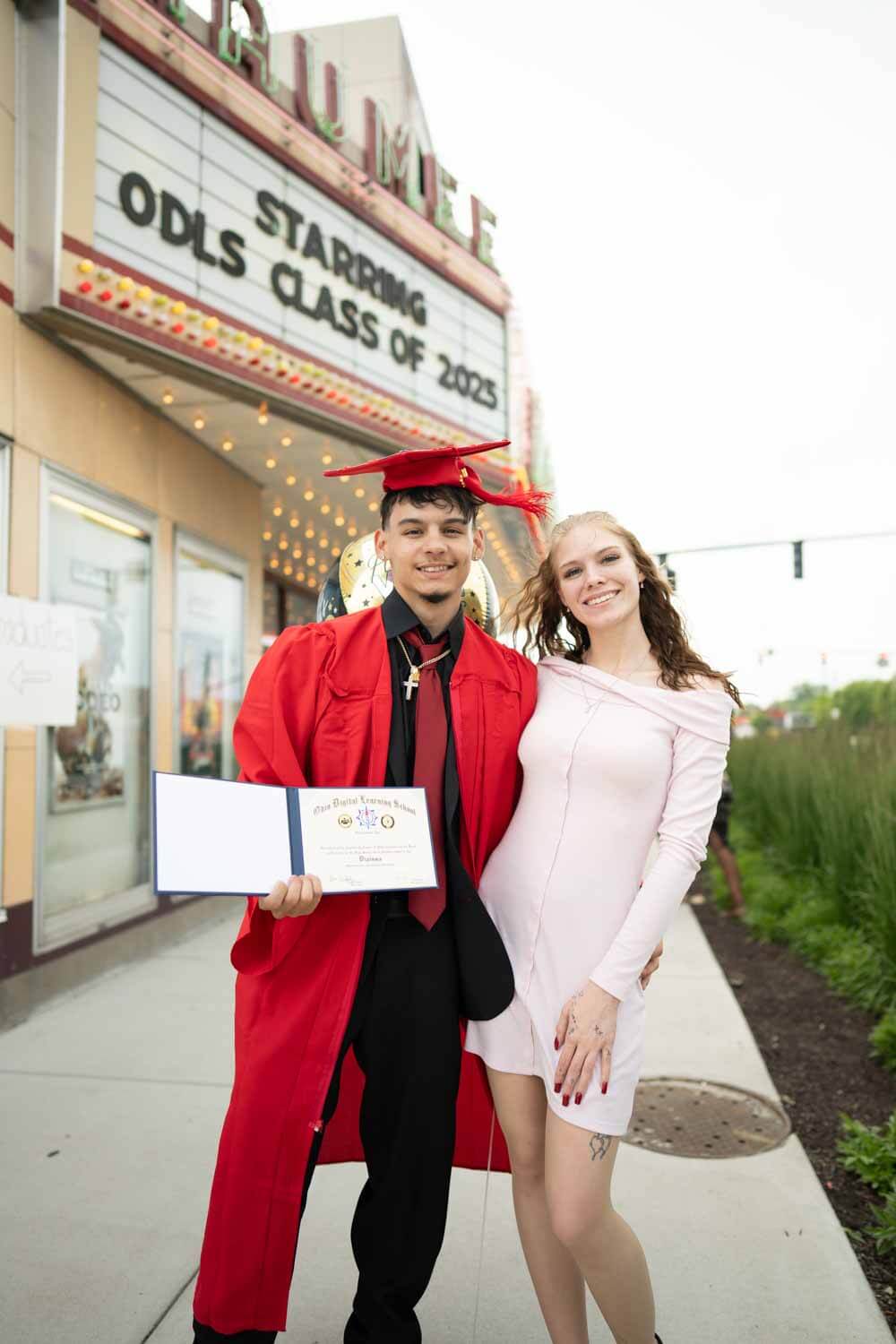 A graduate in a red cap and gown stands smiling with his diploma beside a woman in a light dress.