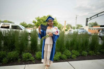 A smiling graduate in a blue cap and gown holds a diploma and a bouquet.