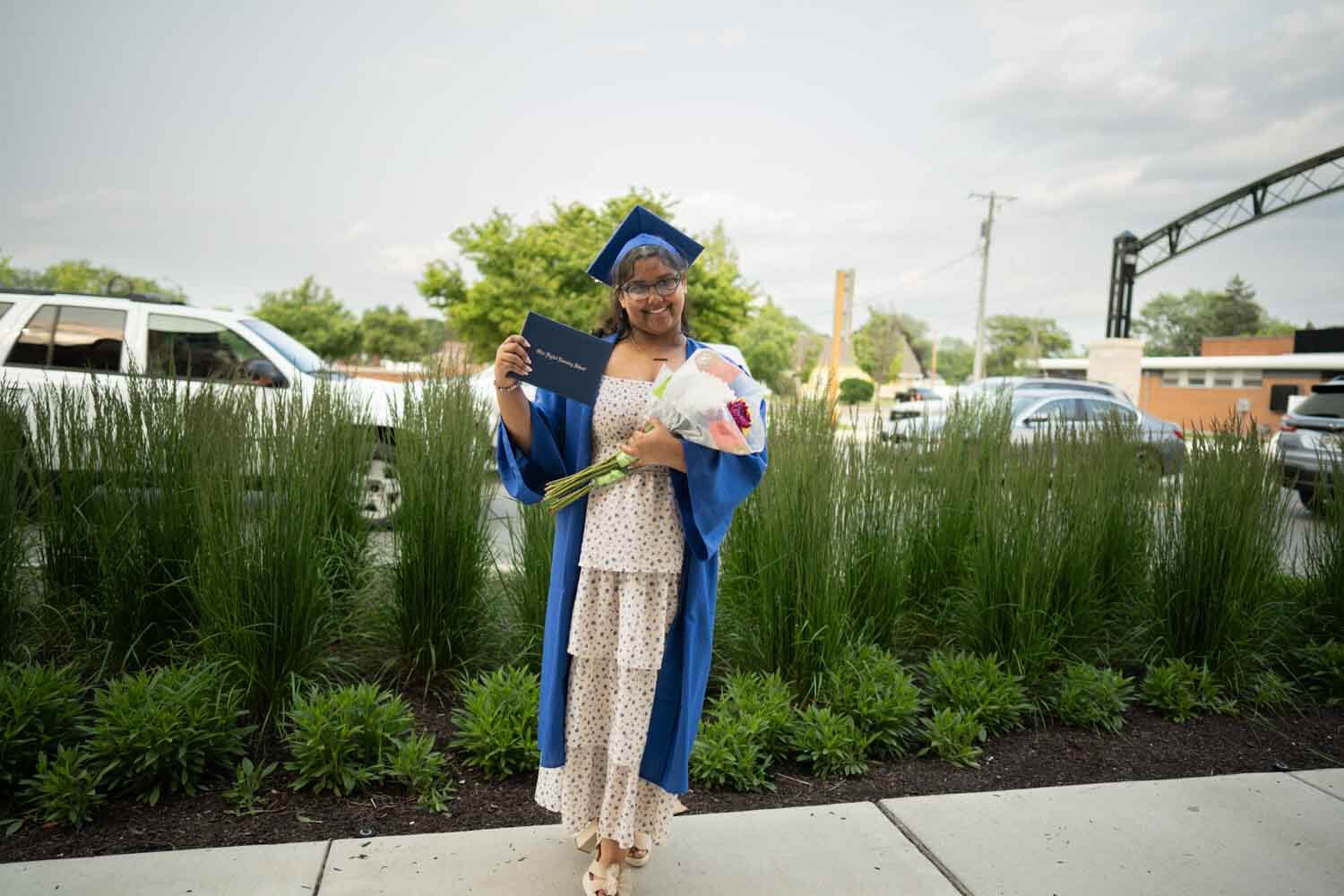 A smiling graduate in a blue cap and gown holds a diploma and a bouquet.